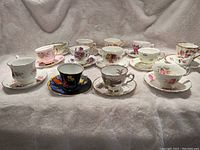 Overview photo of all 13 tea cups and saucers arranged in two rows, showing variety of floral patterns and gold trim on delicate bone china.