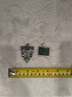 Two silver pendants on white textured fabric surface with measuring tape for scale, showing sizes of 1 to 2 inches