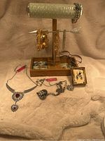 Wooden jewelry stand with beige padded horizontal bar holding assorted gold bangles and bracelets, multiple jewelry pieces on fur background including silver necklaces with red stones and a heart-shaped pendant, earrings, and eyeglasses.