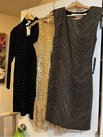 Three ladies' party/cocktail dresses hanging on hangers including a black rhinestone velvet dress, a gold and white beaded dress, and a black dress with silver beading.