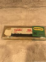 Front view of the N scale Minitrix Carnation Milk hopper car inside original clear plastic box on a textured fabric background.