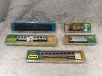 The photo shows five model train pieces in their plastic Minitrix boxes. The items include a yellow locomotive, multiple passenger cars in silver and burgundy colors, and a freight car. The items are arranged on a soft surface.