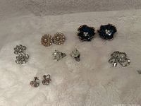 Seven pairs of assorted vintage earrings on white plush surface, showing various styles including pearl, rhinestone, and floral motifs.
