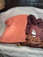 Photo showing two wool blankets laid out on a soft white textured surface. One blanket is rust orange, the other is burgundy with tags visible.