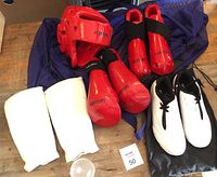 Red helmet, white shin pads, red gloves, red foot pads, white shoes, and blue mesh bag laid out together for overview of entire karate gear set.
