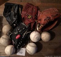 Four left-handed baseball gloves and six baseballs placed on wooden floor, gloves in black, red, and tan colors.
