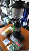 Photo of green Coleman propane lantern with glass globe and black top, sitting on a table with the empty propane canister attached. Background includes various containers and household items.