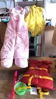 Pink snow pants, yellow pineapple costume, red and yellow life jacket, green bowl with spoons and bib, and pink bubble wand toy on wooden surface.