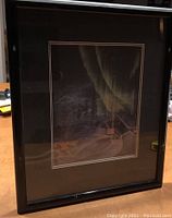 Framed print in dim light showing the northern lights, snow cabin, and mountains, photo taken from a distance with some reflection in glass