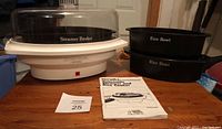 Rival Automatic Steamer and Rice Cooker base unit with steamer basket and dome lid, two black rice bowls, and instruction manual on a wooden surface.