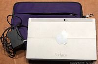 White back of Microsoft Surface tablet showing Surface branding and front camera with included purple case and charger on wooden surface.