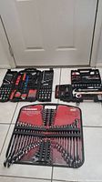 Photo of all three tool sets arranged on a tiled floor showing the Husky wrench set in its red case and two black molded plastic cases with assorted tools.