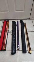 Three pool cues and their carrying cases laid on the floor, showing various lengths and conditions