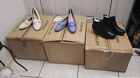 Three pairs of new Studio 1886 ladies shoes displayed on cardboard boxes: beige loafers, blue loafers, and black ankle boots.