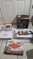 Full lot with boxed Keurig, George Foreman grill, Farberware griddle, copper-infused bacon cooker, and white boxed items on tile floor