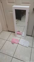 White wall mount cabinet with mirrored door, standing on tiled floor near door. Instruction manual and mounting hardware in pink wrapping placed in front.