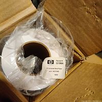 Top view of a roll of HP Universal Bond Paper inside an open cardboard box. The roll is wrapped in plastic and has a label with reorder number Q1396A.