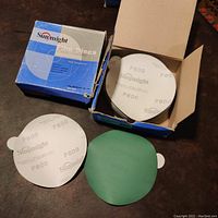 Opened box showing white circular sanding discs marked 'Sunmight' and 'P600'. One disc partially exposed showing both front and back sides, with a tab for handling.