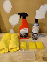 Photo showing orange spray bottle with label 'maxxin detailer', gray refill bottle, two folded yellow microfiber towels, and three yellow clay bars wrapped in plastic in front of a stained wall