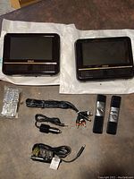 Two RCA 7-inch screen car DVD players laid on white packing cloth with a remote, cables, and straps on a brown surface.