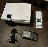 White RCA projector with black lens, RC remote control in plastic wrap, and power adapter on a dark surface.