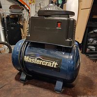 Front side view of blue Mastercraft tabletop air compressor with power switch and logo visible, placed on a dark brown surface with a cluttered background.