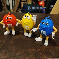 Front view of three M&M's character plastic coin banks in red, yellow, and blue colors with their faces, arms, and legs visible.