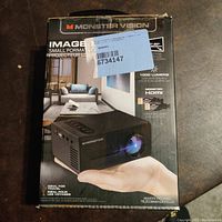 Front side of boxed Monster Vision Image Mini portable projector showing projector image, key specs including 1000 lumens, HDMI support, 30-80" screen size, remote included.