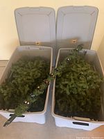 Two gray plastic storage bins open, filled with artificial Christmas tree branches; center pole with colored stickers resting across bins.
