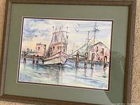 Framed watercolor painting showing two ships at a harbor with wooden docks and background buildings, rendered in soft pastel colors with signature visible.