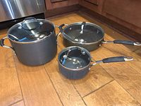Photo showing three Calphalon cooking cookware items including large pot, skillet, and small saucepan, all with glass lids and black handles on wooden floor.