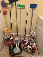 Photo showing multiple cleaning tools with long handles standing vertically. In front are boxes and bottles of cleaning supplies, mop bucket, dustpan and brush, and plastic trays.