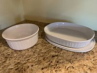 Two vintage CorningWare French White casseroles, oblong dish with lid on right, round dish without lid on left, placed on countertop.