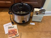 Image of Rival Crock Pot slow cooker next to Rival electric can opener on a wooden surface, with Crock Pot cookbook in front