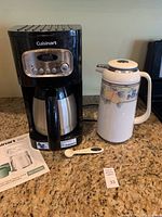 Front view of Cuisinart coffee maker with attached thermal carafe, ceramic Mikasa carafe with floral pattern, and coffee measuring spoon on countertop.