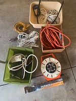 Top-down photo showing coiled electrical extension cords in yellow, orange, brown, and multiple white cords, also beige and white ropes in bins, white surge protector power strip, orange and white cord winder, and packaged Duracell 7 C batteries.
