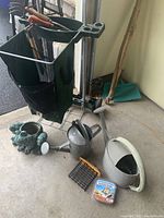 Green fabric wheeled garden tool cart with compartments and mesh bag for clippings, with various garden tools in top tray, watering cans, decorative planter, seed tray and lures placed beside it