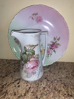 Photo of German hand painted china pitcher positioned in front of a matching plate with floral design.