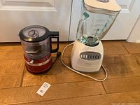 Photo showing both the red KitchenAid food chopper and the white Oster blender side by side on a wooden floor.