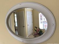 Front view of the white oval mirror hanging on the wall reflecting open door and flower arrangement.