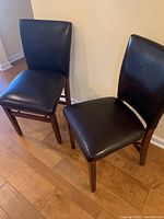 Two dark brown vinyl padded folding chairs with wooden frames standing side by side on wood floor near wall.