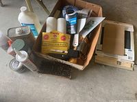 Overview of painting tools and supplies in a cardboard box and surrounding area, showing rollers, brushes, and bottles.