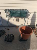 Three black plastic plant dollies on rollers, large terracotta-colored plastic pot, and green metal plant stand with planter shown outside on concrete against siding.