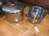 Shows full set including large stock pot with lid and the double boiler with built-in strainer insert and lid on wooden floor.