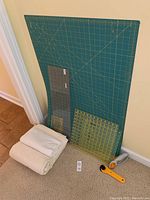 Photo showing large green OLFA quilting rotary cutting mat leaning against wall, two acrylic measuring rulers in front, two pieces of fabric folded and laid on the floor, and two rotary cutters on the floor.