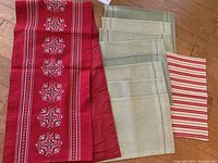 Red Christmas table runner with white cross-stitched snowflake patterns and green accents, alongside eight beige placemats and one red and white striped placemat on wooden floor.