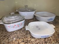 Four vintage Corning Ware casserole dishes with two interchangeable glass lids. One dish has Rosemarie Tulip pattern, two have Cornflower Blue pattern, and one plain white.