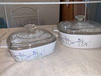 Two vintage CorningWare Shadow Iris casserole dishes of different sizes with clear glass covers, side view.