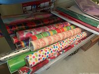 Multiple partial and full rolls of wrapping paper with different designs on a shelf, some rolls partially used, several colors and patterns visible including polka dots and musical notes.