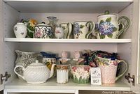 Shelf with multiple pitchers, teapots, and decorative ceramic items, showcasing floral and fruit designs on earthenware pieces.